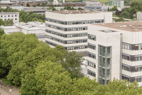 BCI: Rechts im Bild befinden sich zwei hohe graue Gebäude mit vielen Fenstern der Fakultät Bio- und Chemieingenieurwesen. Davor und weiter links im Bild Bäume.